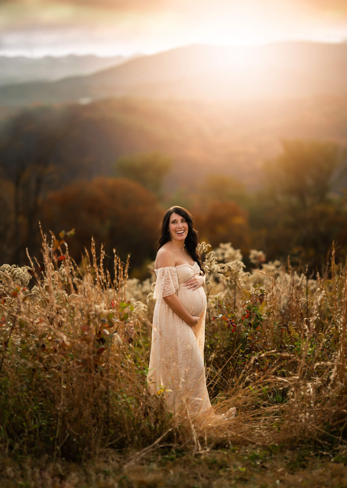 A beautiful mama to be in a long lace gown laughs while cradling her baby bump