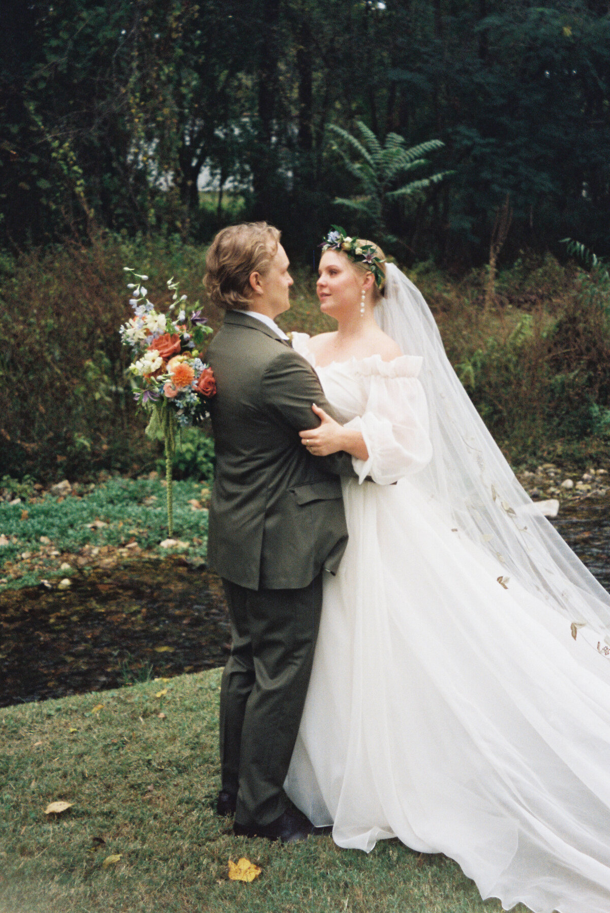 Wedding couple embracing outdoors beside a stream, with the bride holding a lush garden-style bouquet featuring roses, delphinium, dahlias, and greenery, wearing a romantic veil adorned with botanical appliqué.