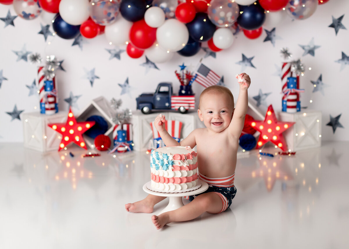 America Patriotic themed cake smash. Baby boy wearing flag-print short is sitting in front of a flag decorated cake. His hands are in the air full of icing and smiling a the camera. In the background, there are red, white, and blue balloons, silver stars, American flags, and a vintage blue truck.
