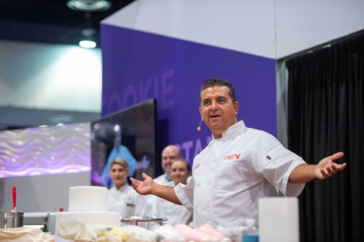 Cake Boss speaking with white undecorated cake in front of him at IBIE