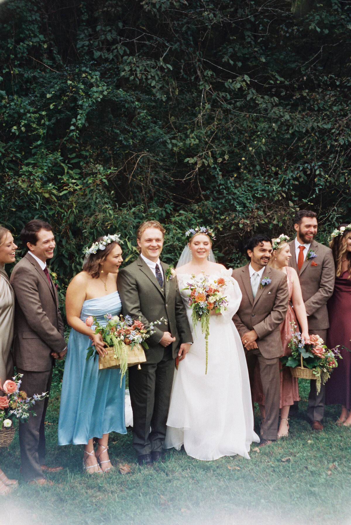 Film photograph of the wedding party standing together outdoors with bridesmaids holding floral baskets and the bride holding a colorful garden-style bouquet. Designed by a Northwest Arkansas wedding florist for a whimsical outdoor celebration.