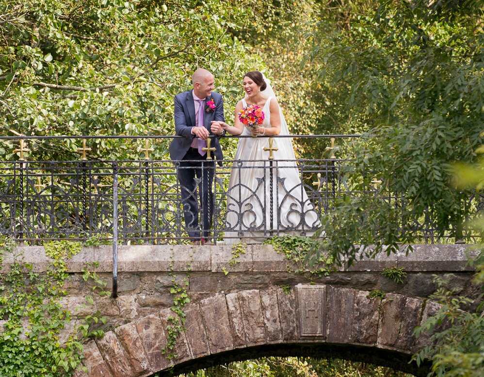 Bride with black hair, wearing a princess wedding dress and holding a colourful bouquet, standing on an old bridge with her groom wearing a blue suit and purple tie, surrounded by trees in Killarney National Park