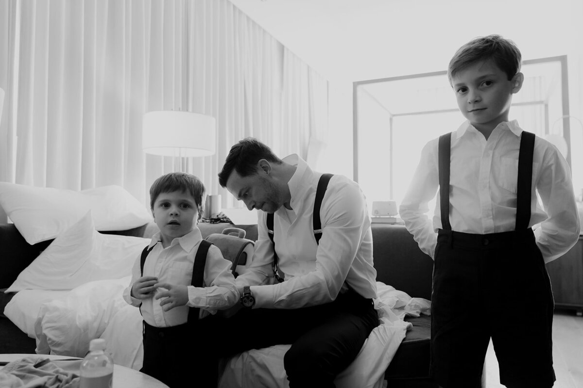 Groom helps child to put on suspenders . Groom sits on bed, both boys look at the camera.