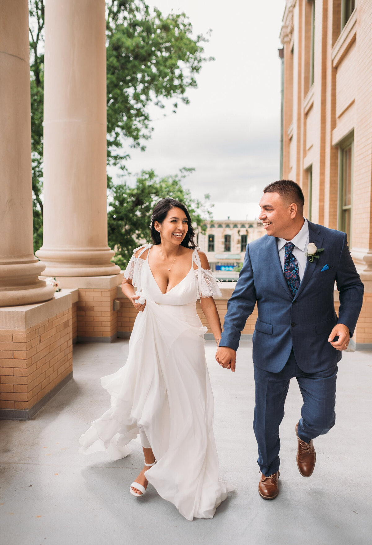 Couples Photography, woman in a  wedding dress is holding hands with man in a  suit, running towards the camera with smiles on their faces.