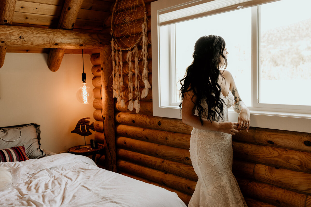 bride in a lace wedding dress standing in front of a cabin window