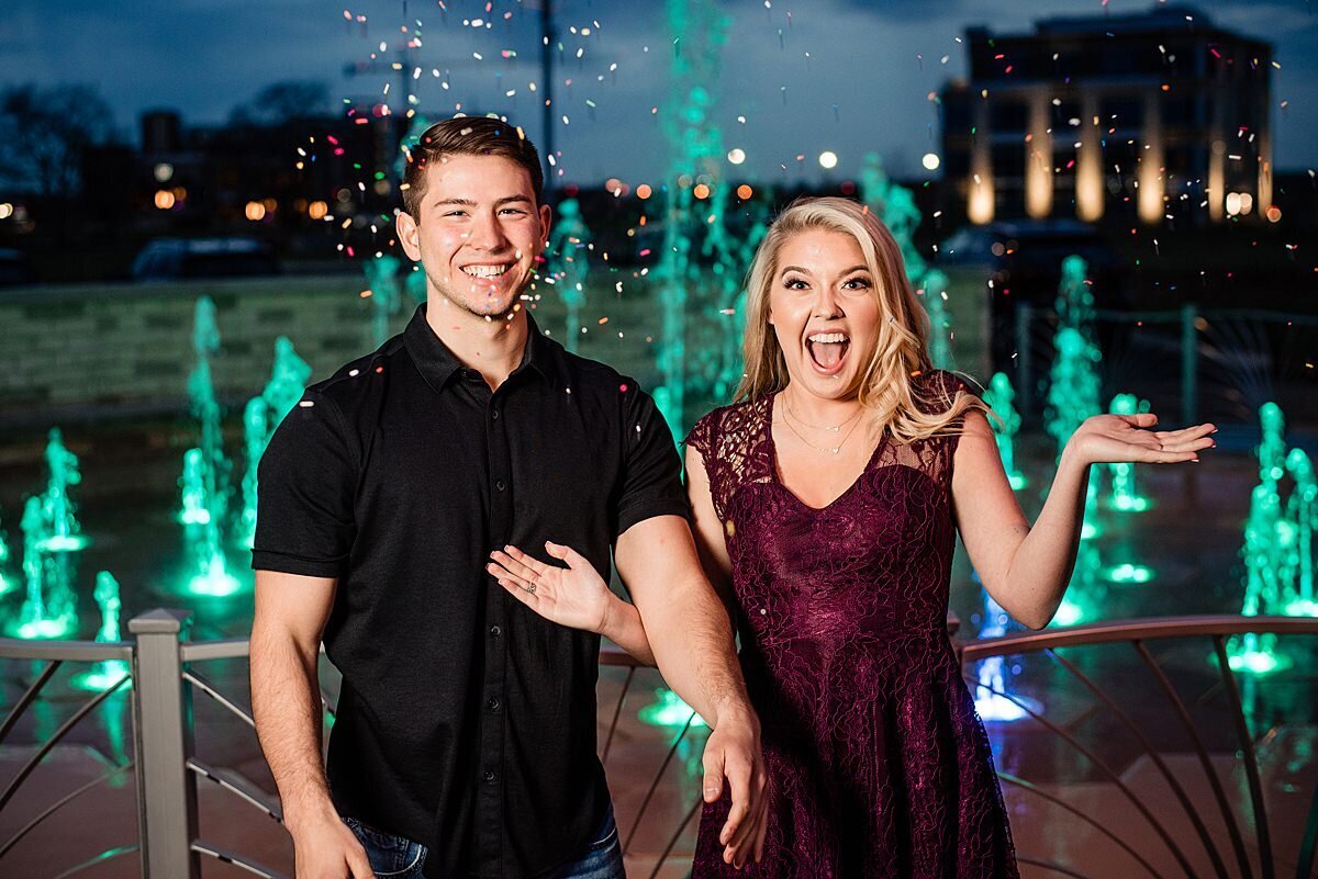 The groom stands next to the bride with his arms at his side. He is wearing a black button down shirt. The bride has her hands thrown in the air as she tosses ice cream sprinkles in the air. She is wearing a maroon sleeveless lace dress. They are standing in front of a fountain lit up with teal blue lights.