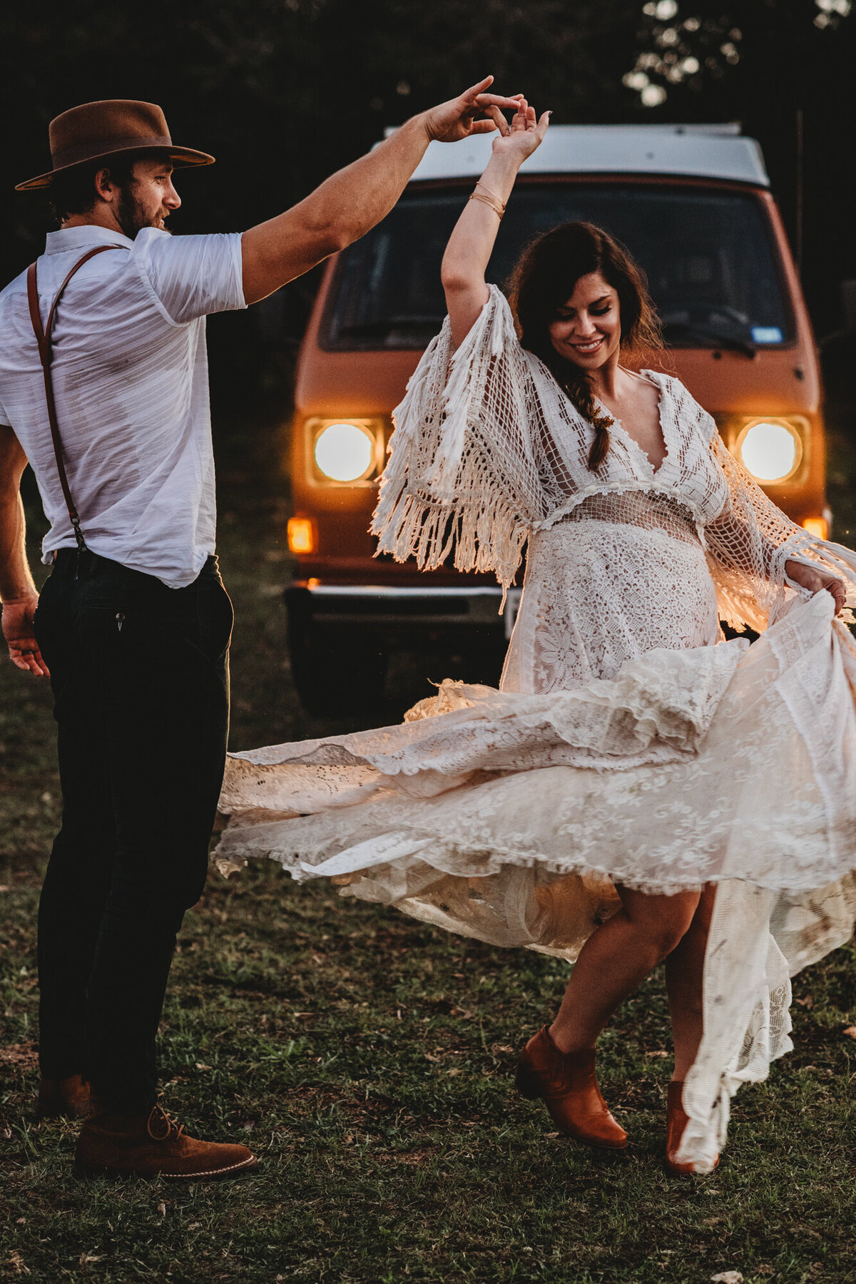 Couples Photography, Man in suspenders and a brown hat holding the hands of a woman in a white lace wedding dress, twirling her in front of a rust colored vintage VW bus.