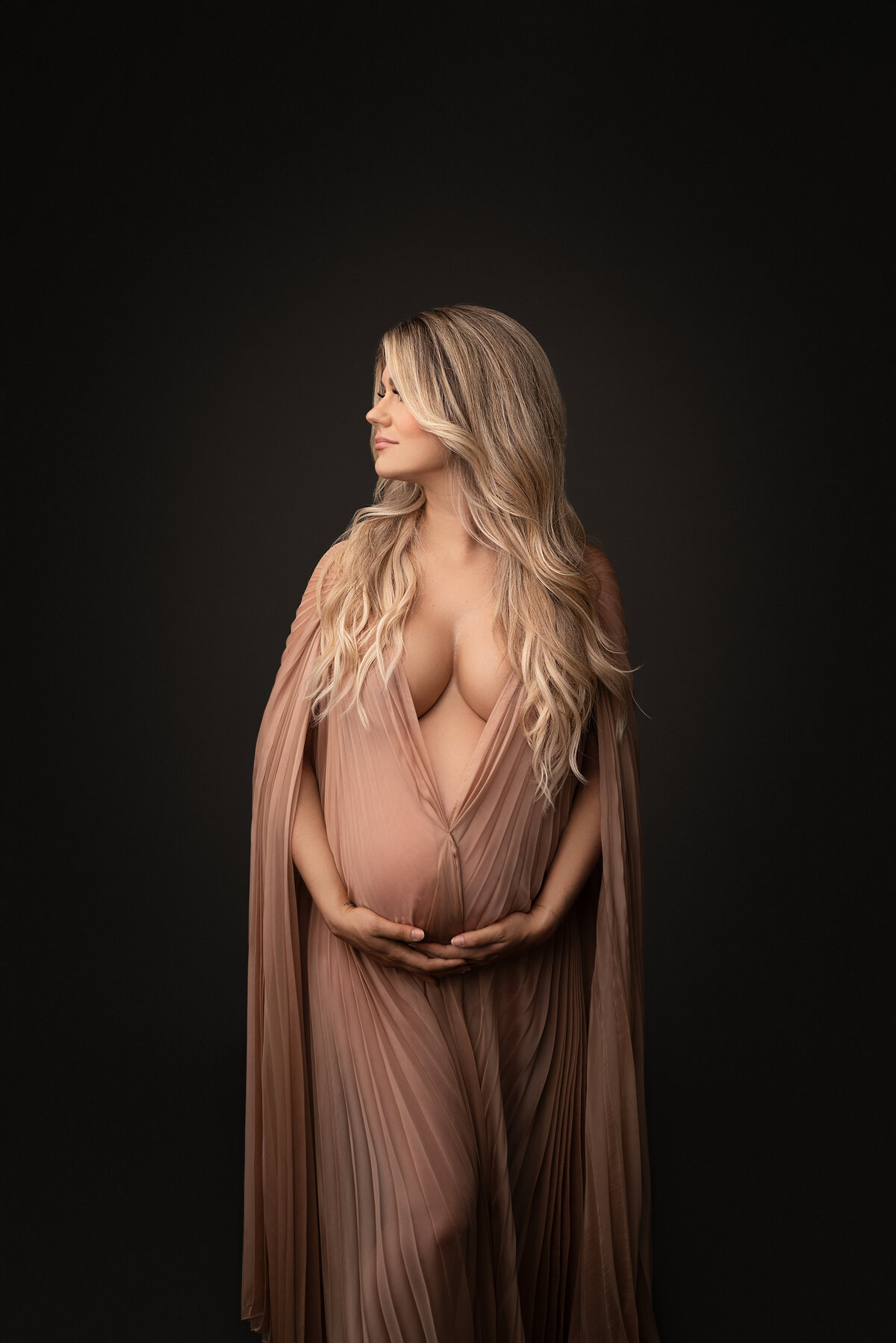 Philadelphia Main Line's best maternity photographer Katie Marshall captures expectant mom for a fine art maternity photoshoot. Expectant mom in a floor length dusty rose maternity photoshoot dress with caped sleeves has her body facing the camera. Her hands are gently resting underneath her baby bump. She is looking over her shoulder looking toward the light.