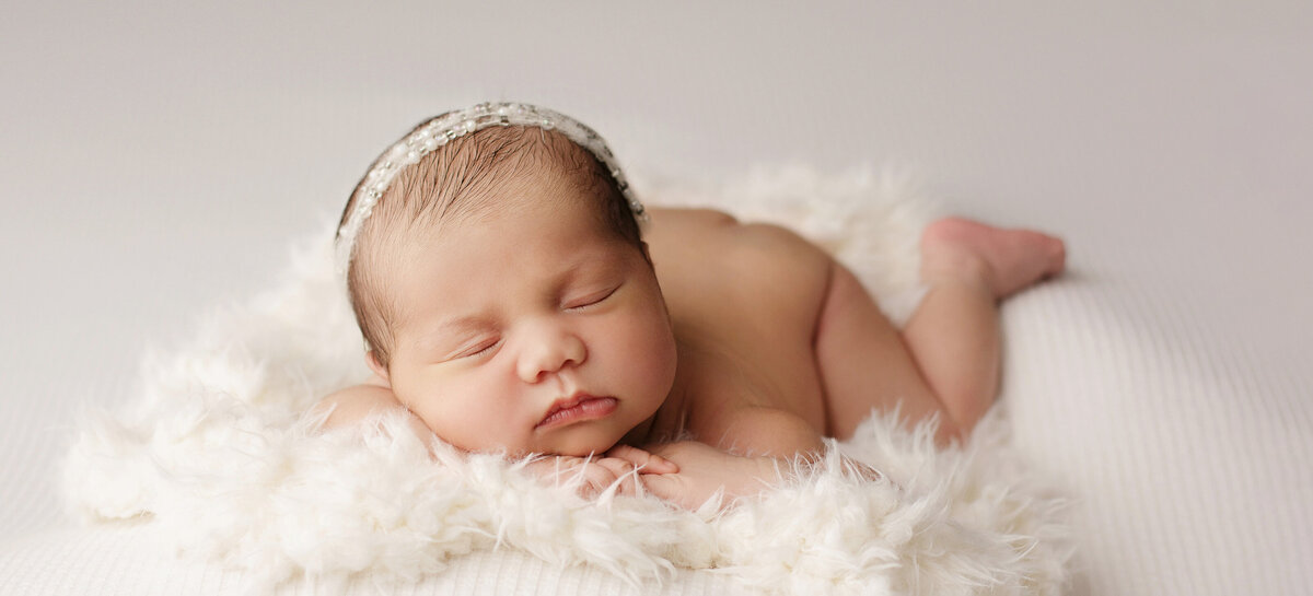 Newborn girl posed on a soft furry white texture facing forward with her head resting on her hands.