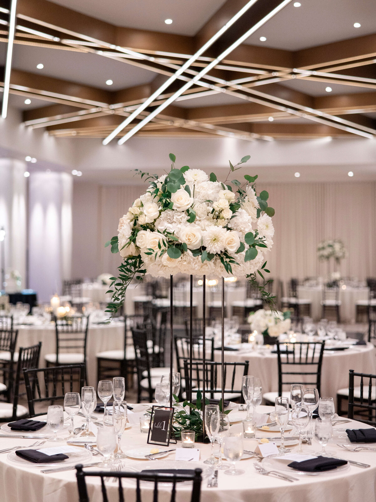 Elegant ballroom with tables set for an event. Each table features black chairs, neatly arranged glassware, and a centerpiece of white roses and greenery on a tall stand. Soft lighting highlights the modern geometric ceiling design.