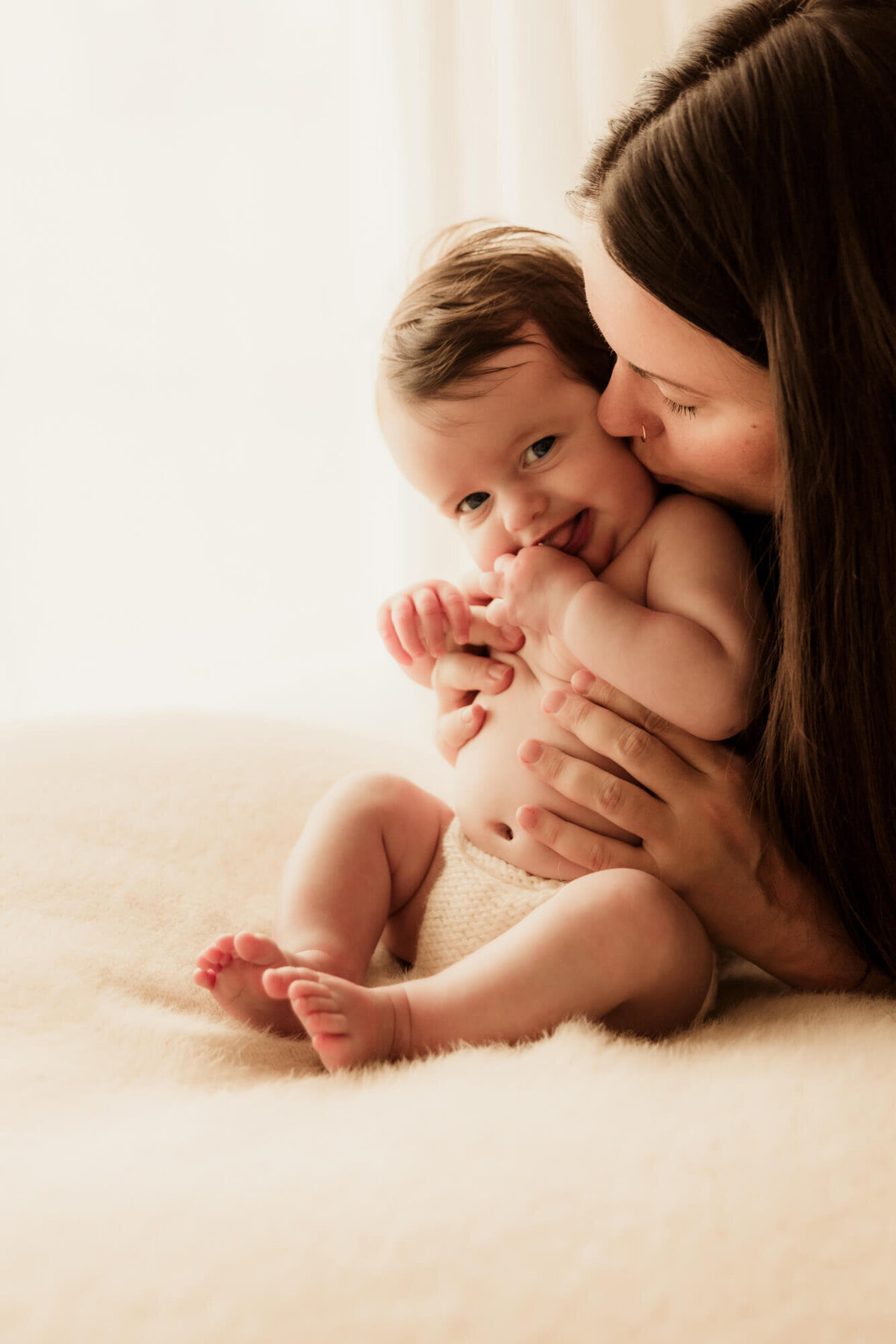 Baby girl smiles with her tongue out as her mother kisses her on the cheek.