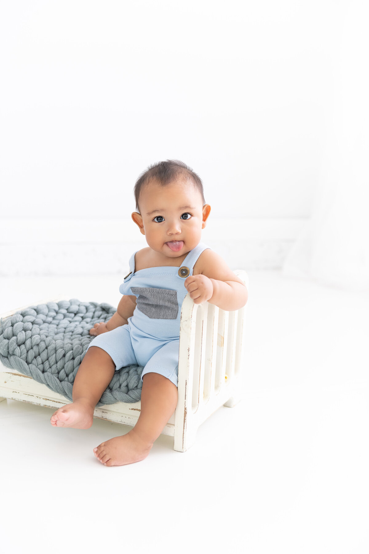 A young toddler boy in blue overalls sits on a small wooden bed with tongue out