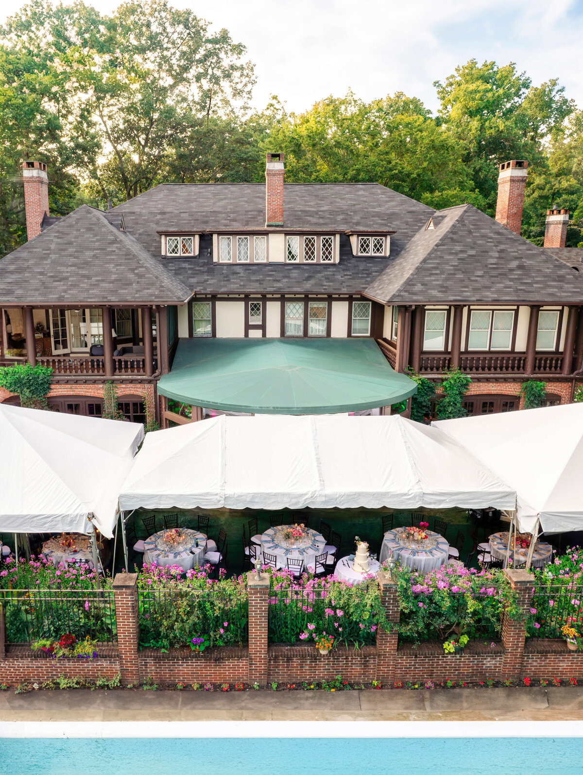 A large, elegant house is surrounded by lush greenery. White tents are set up in the garden, with tables and chairs visible underneath. Colorful flowers line the brick and wrought-iron fence. The sky is partly cloudy.