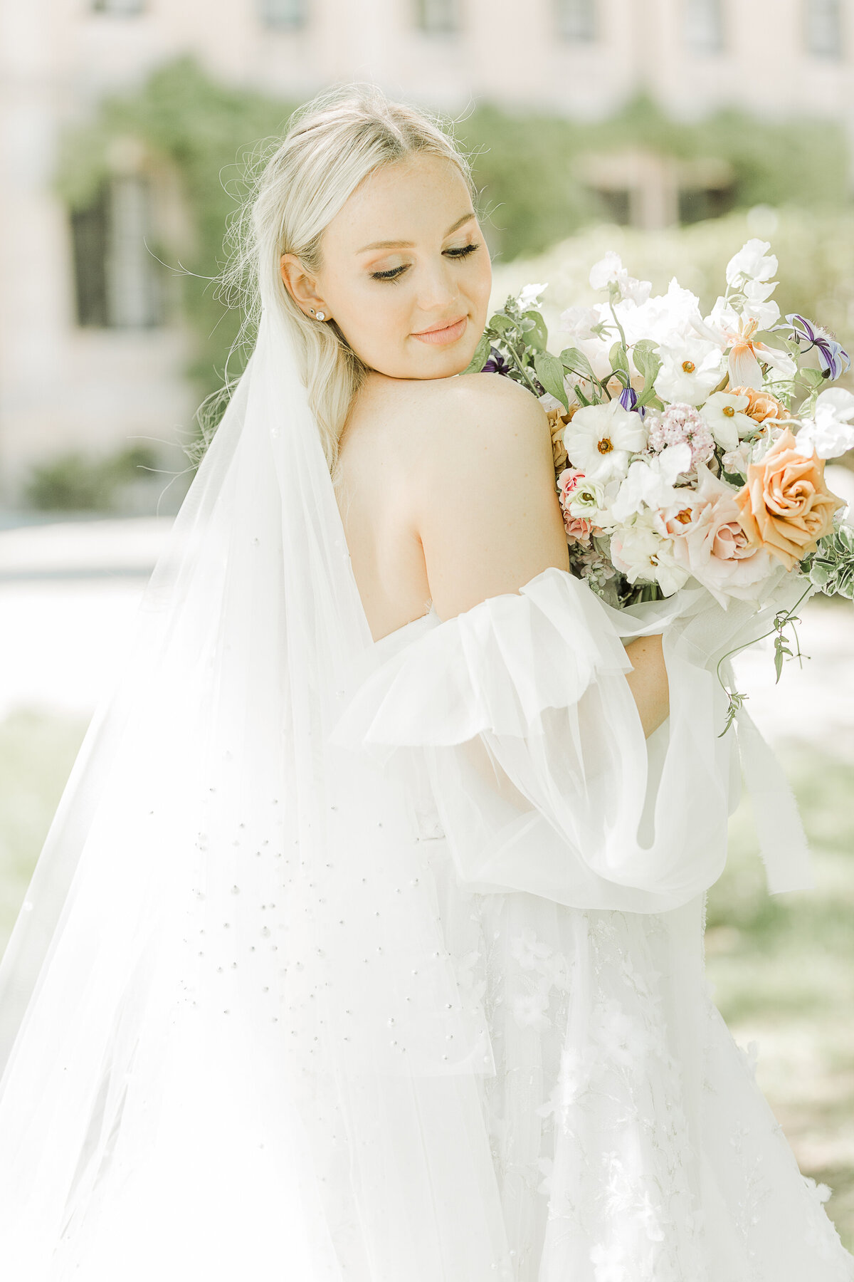 Editorial bridal image captured by Lia Rose Weddings. A beautiful bride in an off-the shoulder gown is looking over her shoulder and holding her bouquet under her chin.