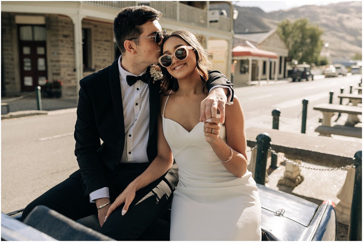 bride and groom sitting on convertible car in queenstown with sunglasses on editorial style holding hands in historic town