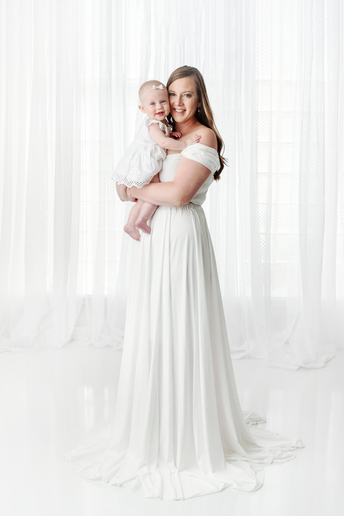 st-louis-motherhood-photographer-mother-holding-baby-both-smiling-at-camera-wearing-monocromatic-white-growns-against-white-background