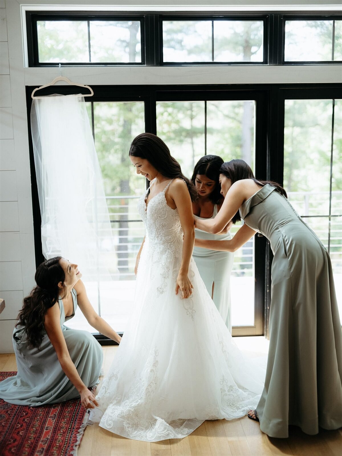 Three bridesmaids in floor-length sage green dresses help bride button and fluff her dress in the modern bridal suite with lots of windows, overlooking wooded area.