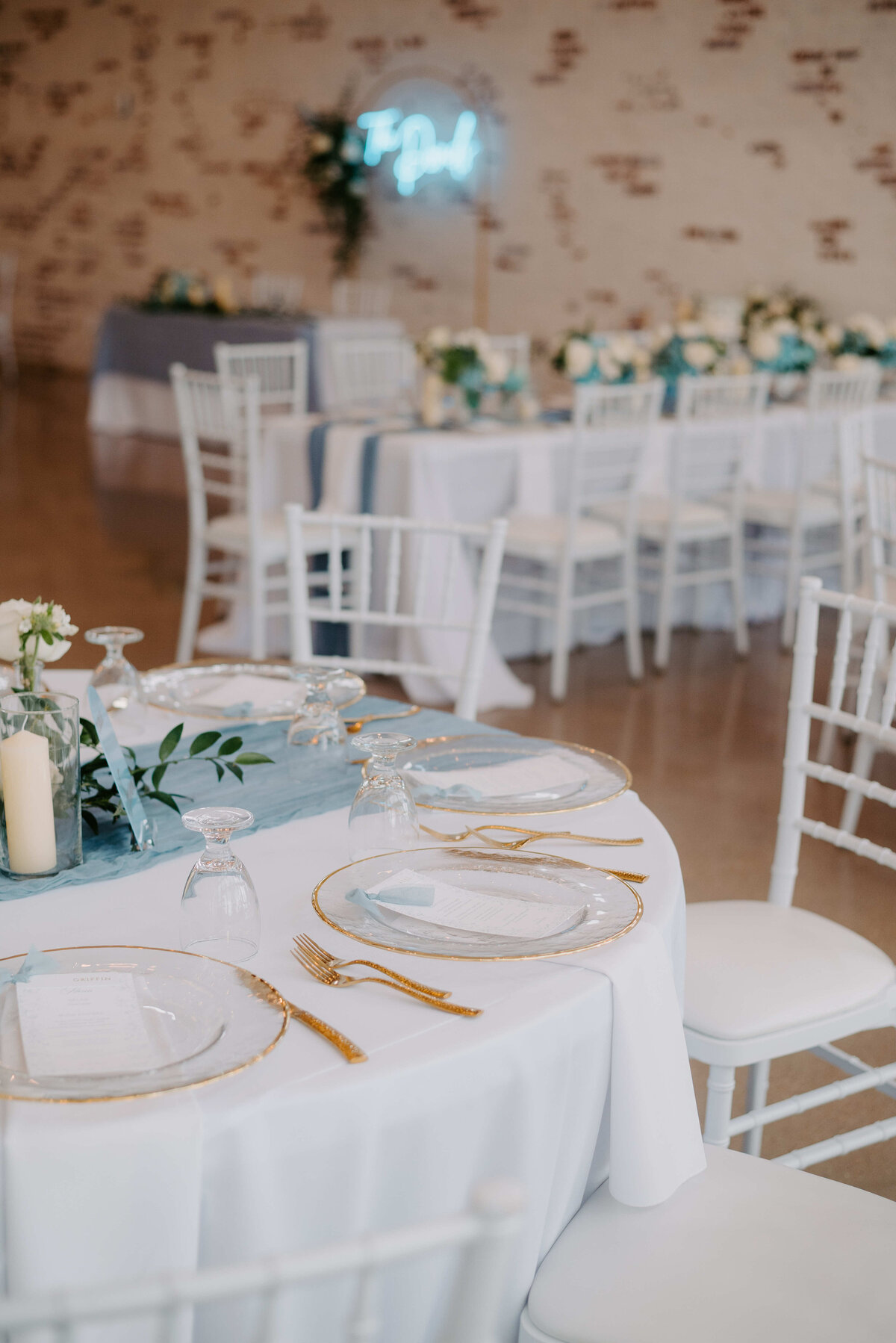 Elegant wedding reception setup with white tables and chairs, gold-rimmed plates, candles, and greenery. A subtle, romantic ambiance is created indoors.