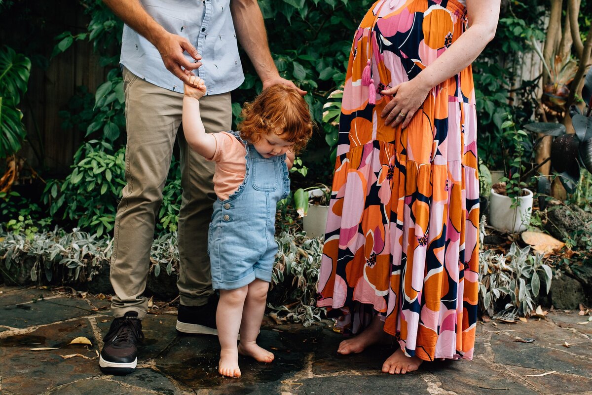 Girl with red hair holding Dad's hands, Mum with pregnant belly in floral skirl.