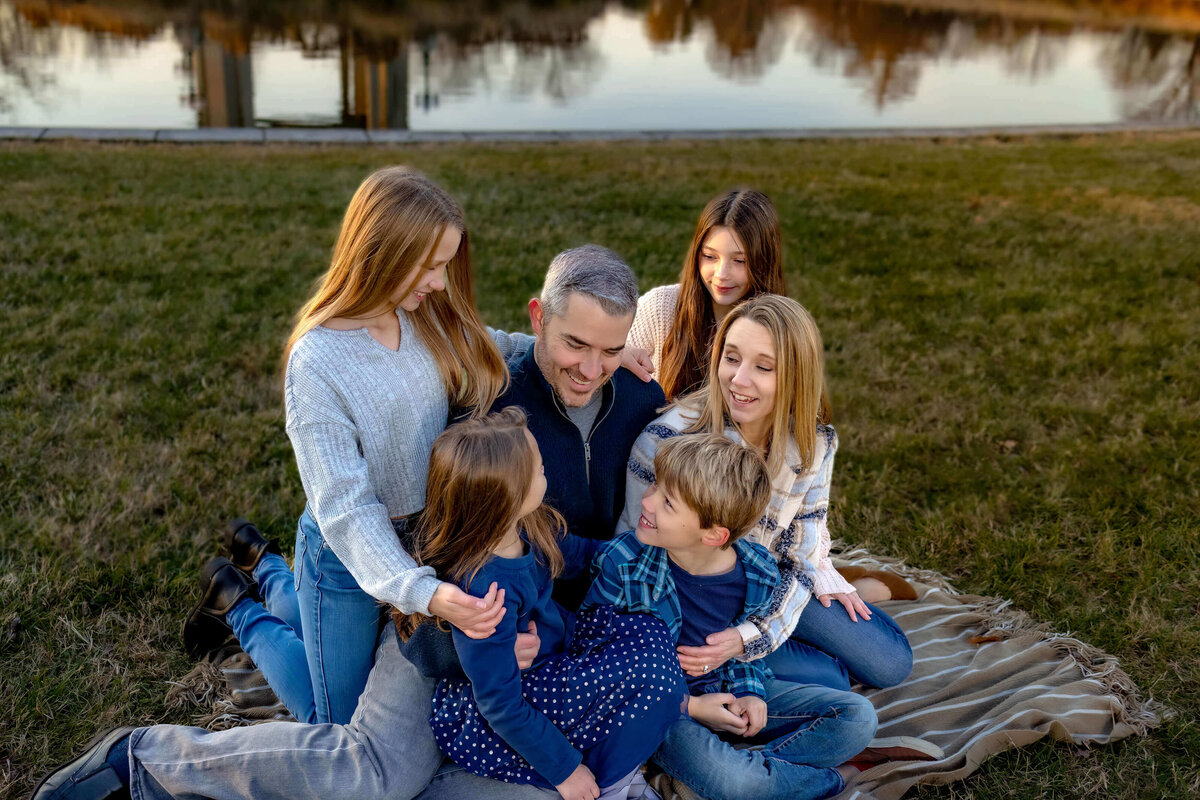 Family of 6 sitting on a blanket in a park by a pond at sunset, everyone is looking at  each other