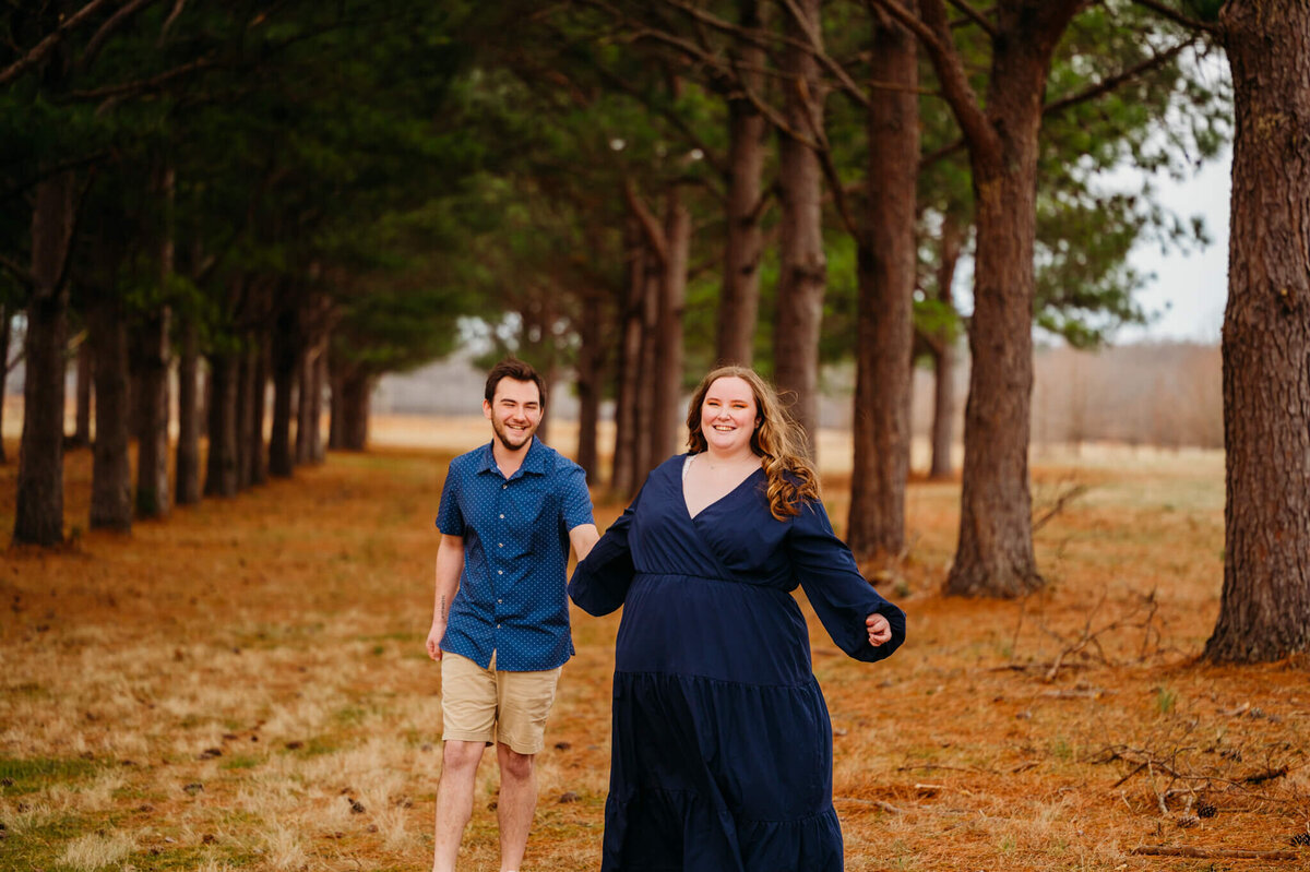 photo of a woman holding her fiance's hand and pulling him through the woods with a happy smile
