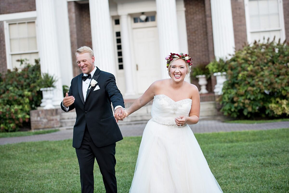 The bride and groom get silly for the camera. Standing in front of a stately mansion, the groom is wearing a black tuxedo with a white shirt and black bowtie. The bride is wearing a strapless dress with a sweetheart neckline and a flowing floor length skirt. The bride has a white, ivory and pink flower crown on her head.