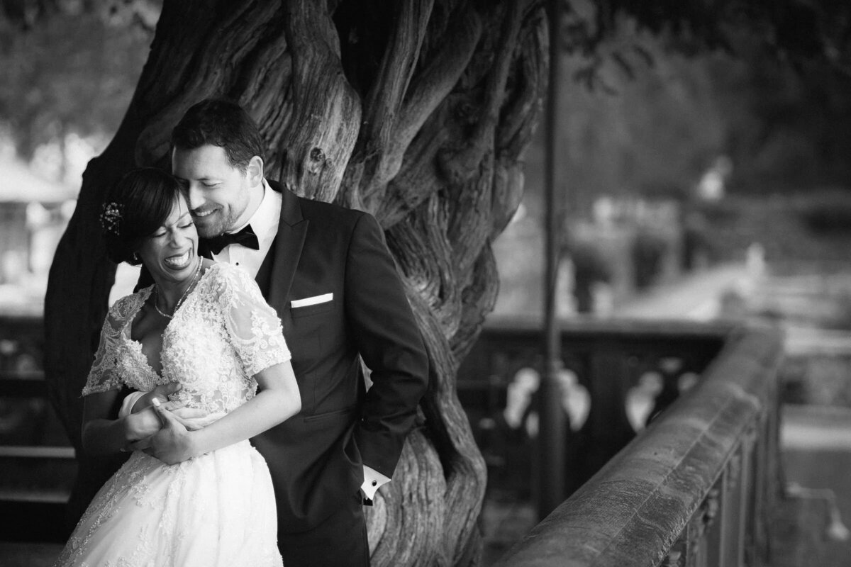 Intimate black and white photo of a bride and groom embracing by an old tree near a waterfront