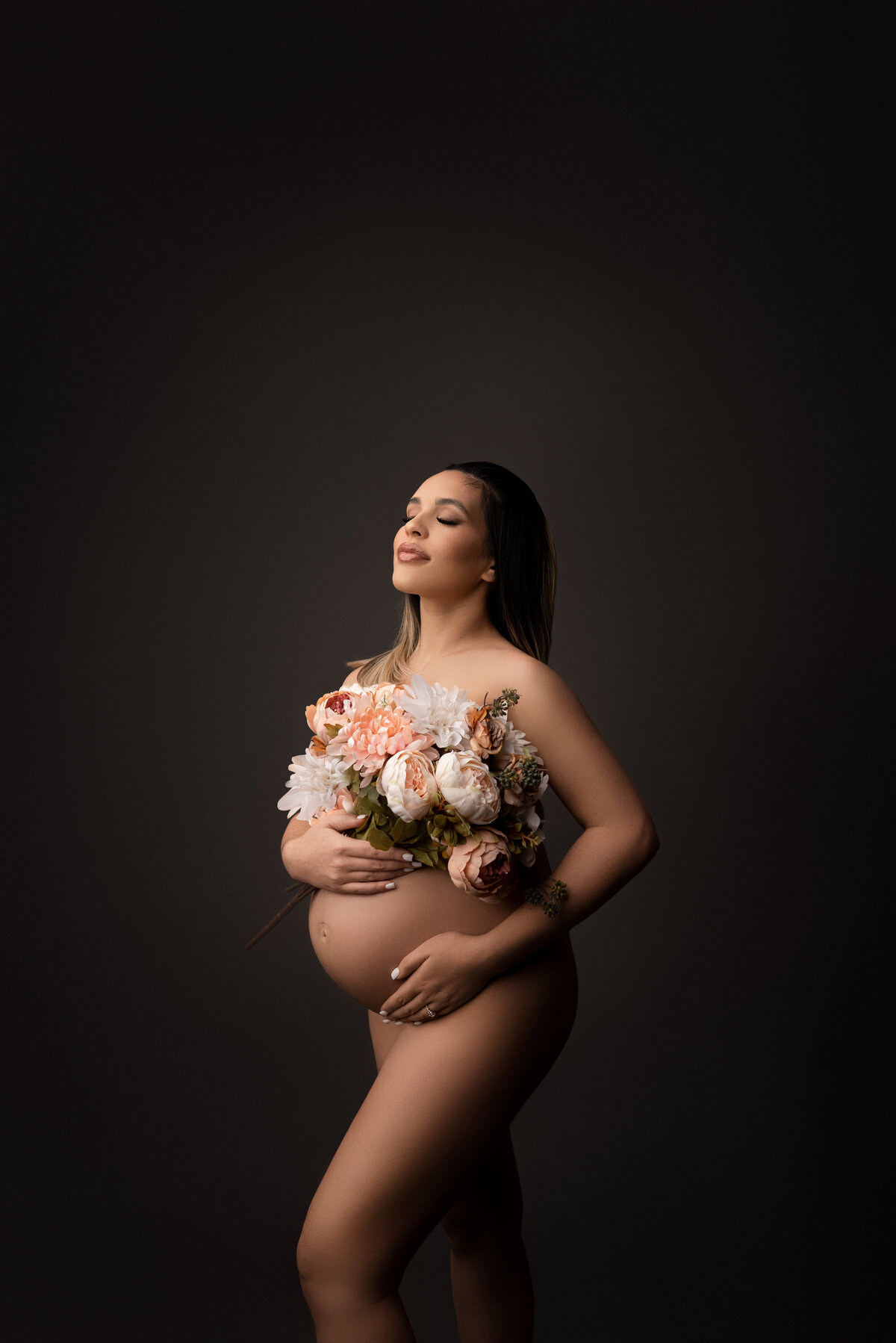 Maternity photograph by Katie Marshall,  premier maternity photographer in New Jersey. The image features a woman standing bare against a grey backdrop, with one leg gently bent and her forearm supporting her baby bump. Her bosom is tastefully covered with a floral bouquet. Her closed eyes are directed upwards towards the light source, creating a play of shadows for added depth and drama, resulting in an artistic and emotive maternity image.