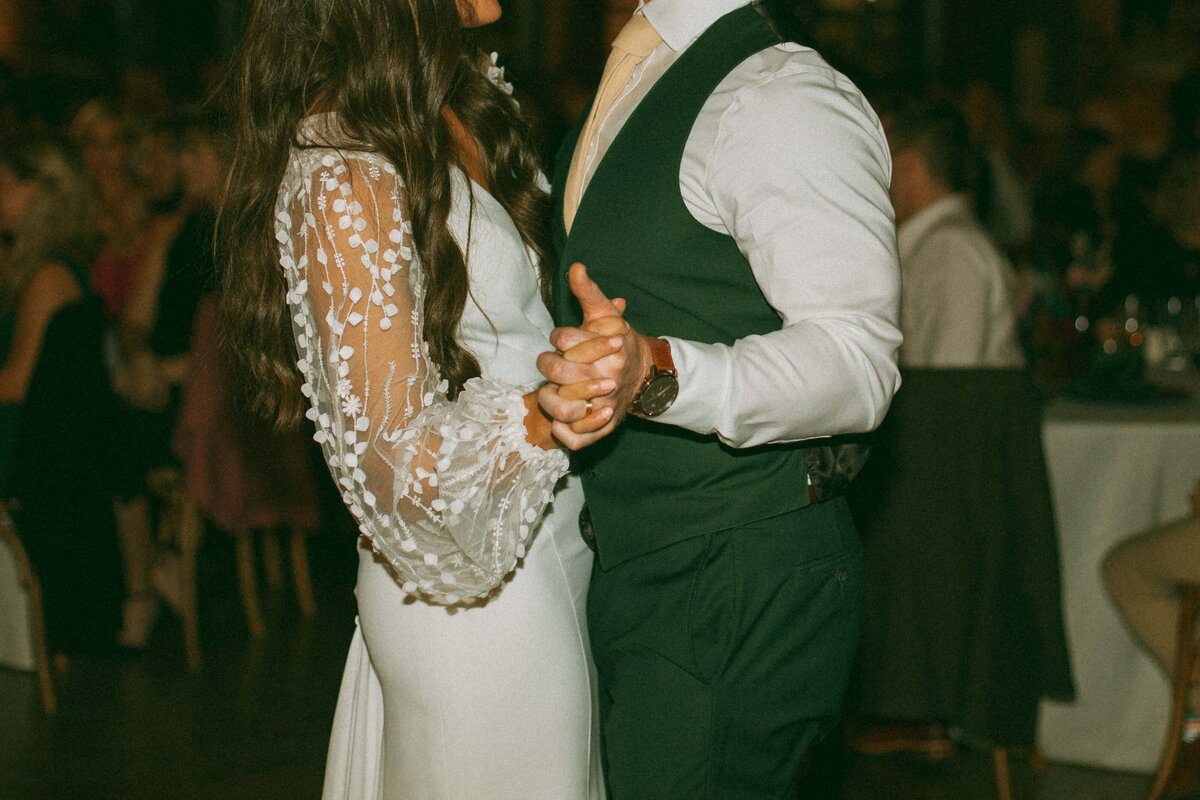 A bride in a lace dress and a groom in a vest and dress shirt hold hands during a dance at an Iowa wedding reception.