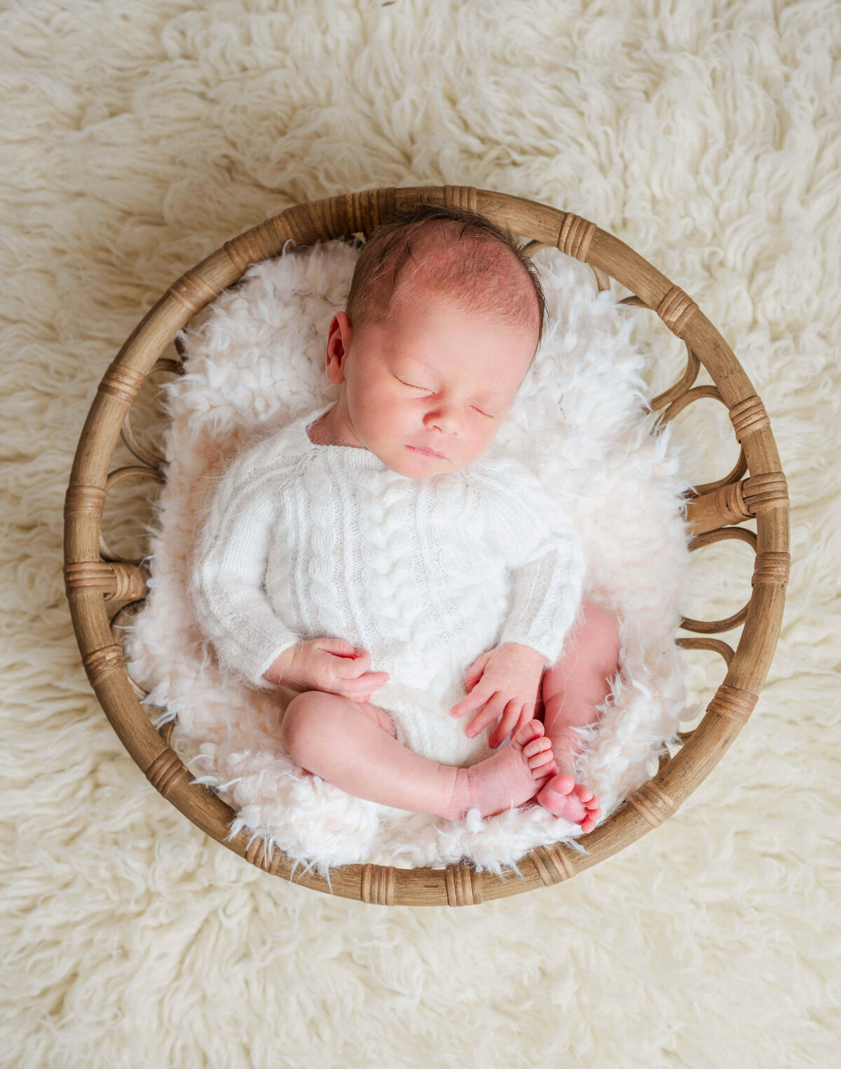 A newborn baby in a  whimsical basket , laying on fuzzy white blanket and wearing a fuzzy sweater.