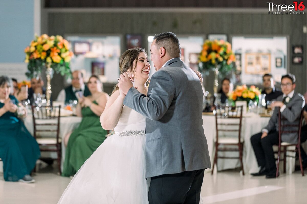 Bride laughs during her first dance with her Groom with wedding guests looking on