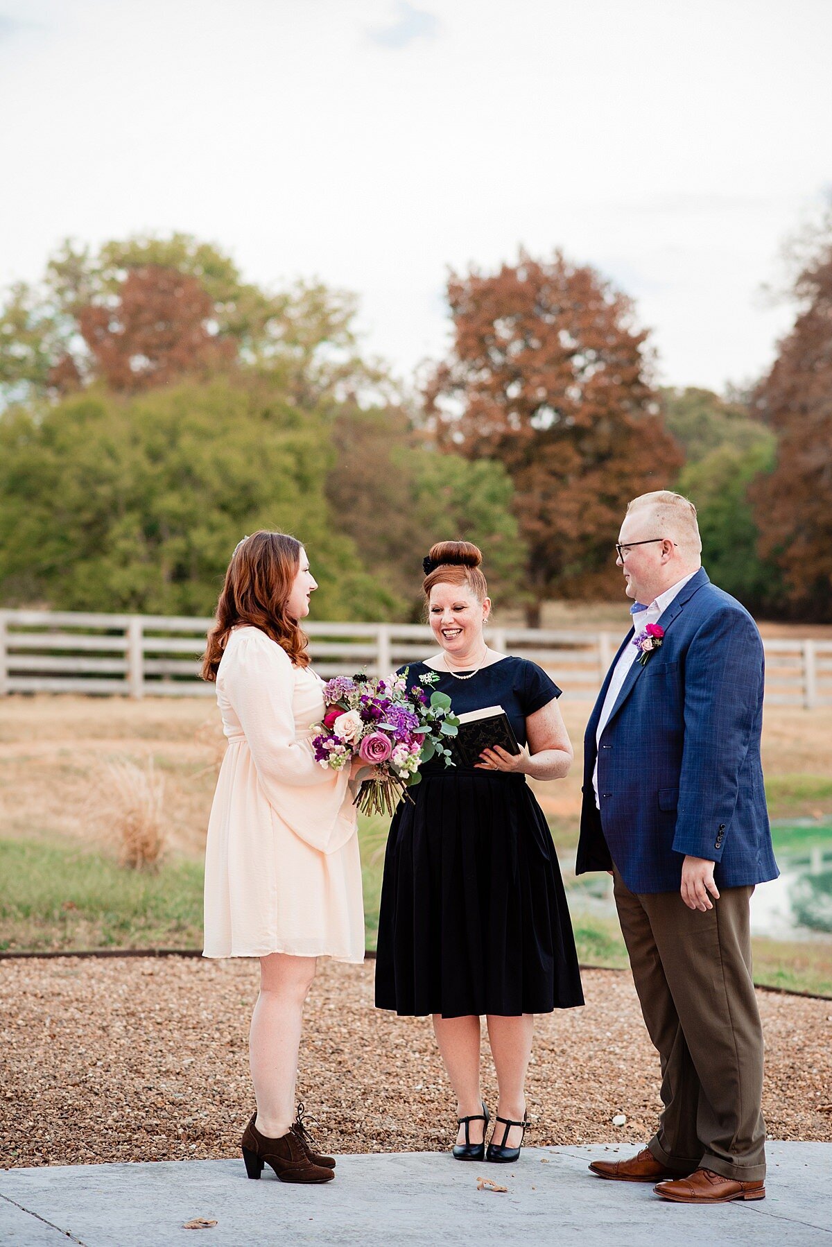 An elopement at Steel Magnolia Barn. The bride is wearing a knee length ivory boho dress with long bell sleeves and short brown booties with heels. She is holding a large bouquet of pink, purple, ivory and burgundy flowers with greenery as she and the groom look at the officiant. The officiant is wearing a knee length black dress t-strap heels. She is holding a leather bound book as she reads. The groom is wearing brown pants and shoes with a white shirt and cobalt blue jacket. He has a pink boutonniere pinned to his lapel.