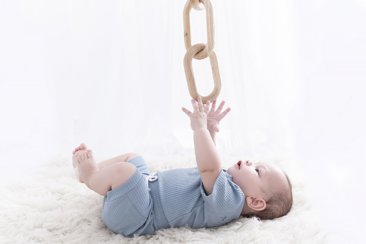 A young toddler boy lays on a bed reaching for a wooden chain link above him in a studio of a childrens photographer Atlanta