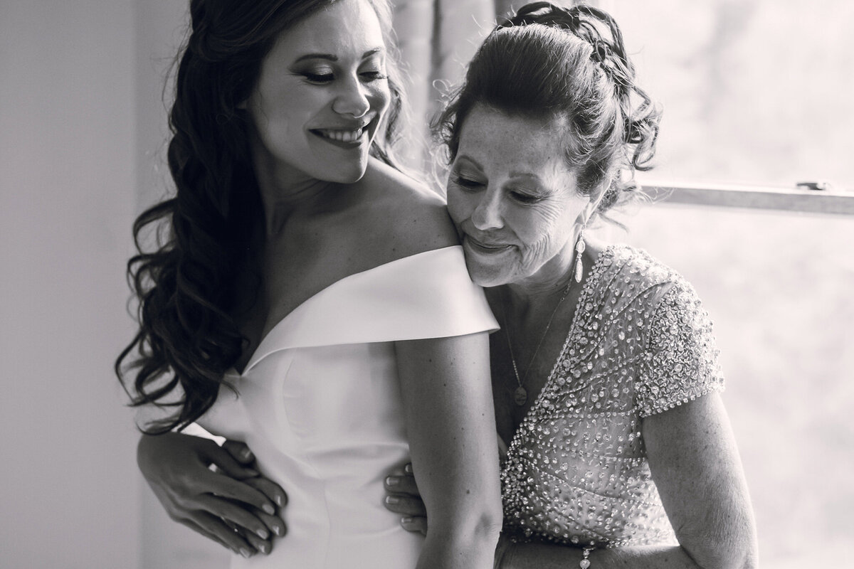 A bride and her mother hugging in front of a window.