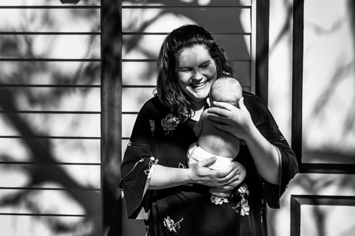 black and white photo of a mom holding newborn son in shadow of trees