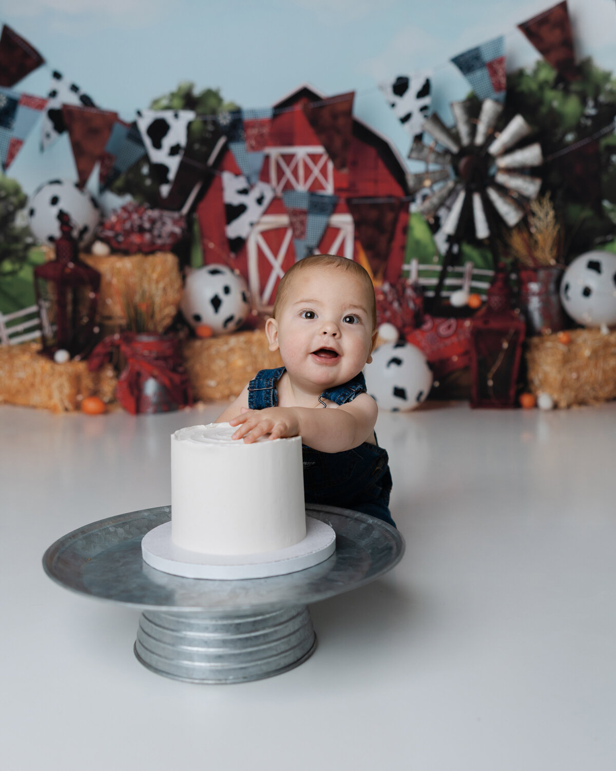 Baby boy enjoying a messy cake smash in front of a red barn backdrop with rustic farm props. Photograph by Yucaipa cake smash photographer Kaitlyn Dawn Photography.