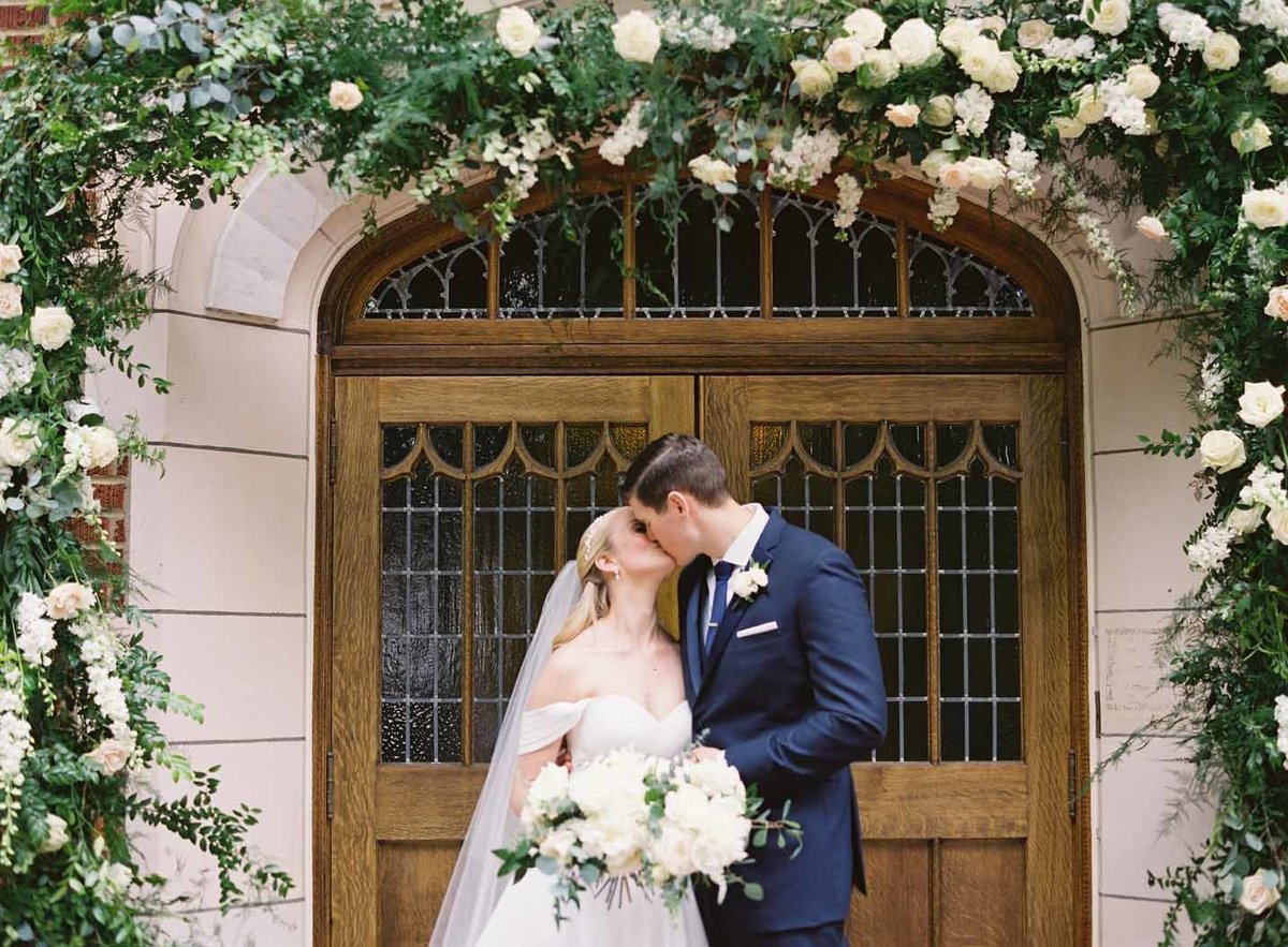 bridee and groom kissing under their floral and greenery arch at the entrance of Villa Academy Chapel