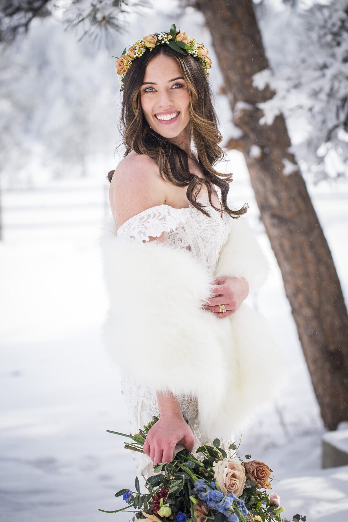 Bride stands in the snow, wrapping her fur around her arms and smiling at the camera.