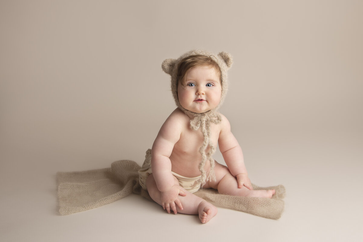 Smiling six month old baby girl wearing a knitted teddy bonnet and sitting on a knitted beige wrap.