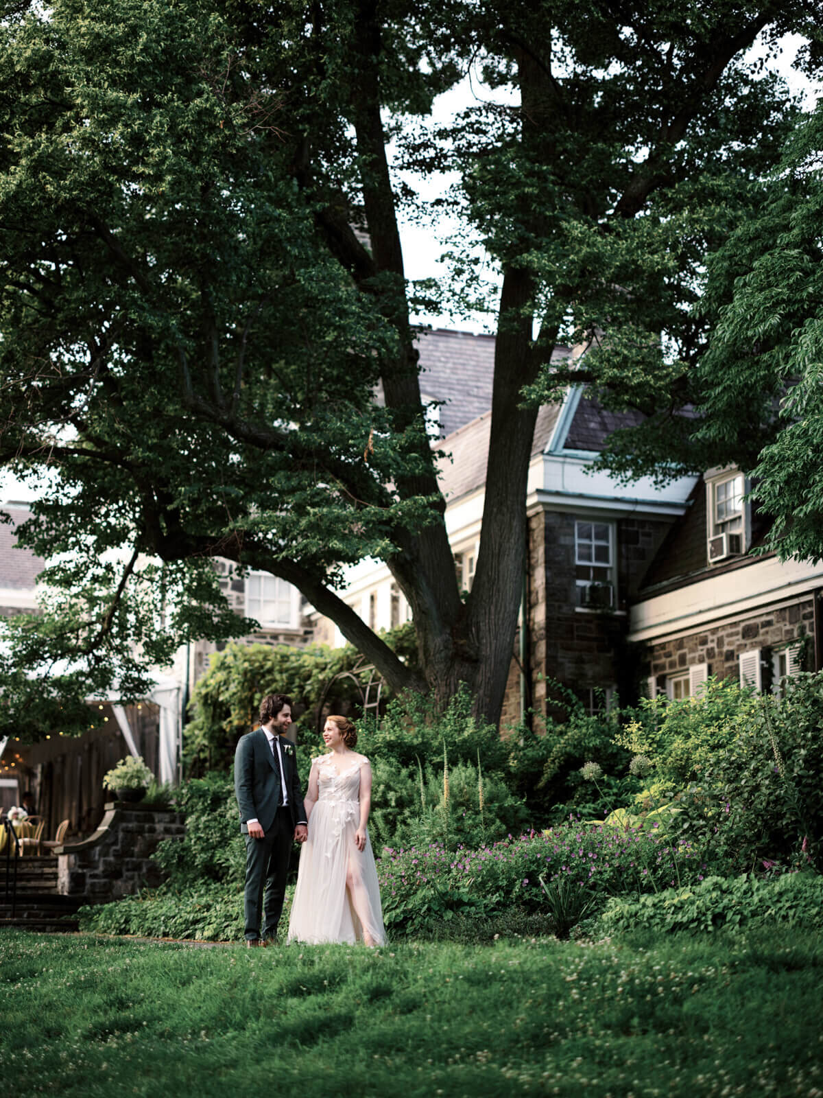 The bride and the groom are standing on a ground covered with grass, with a huge tree and a house on their back.