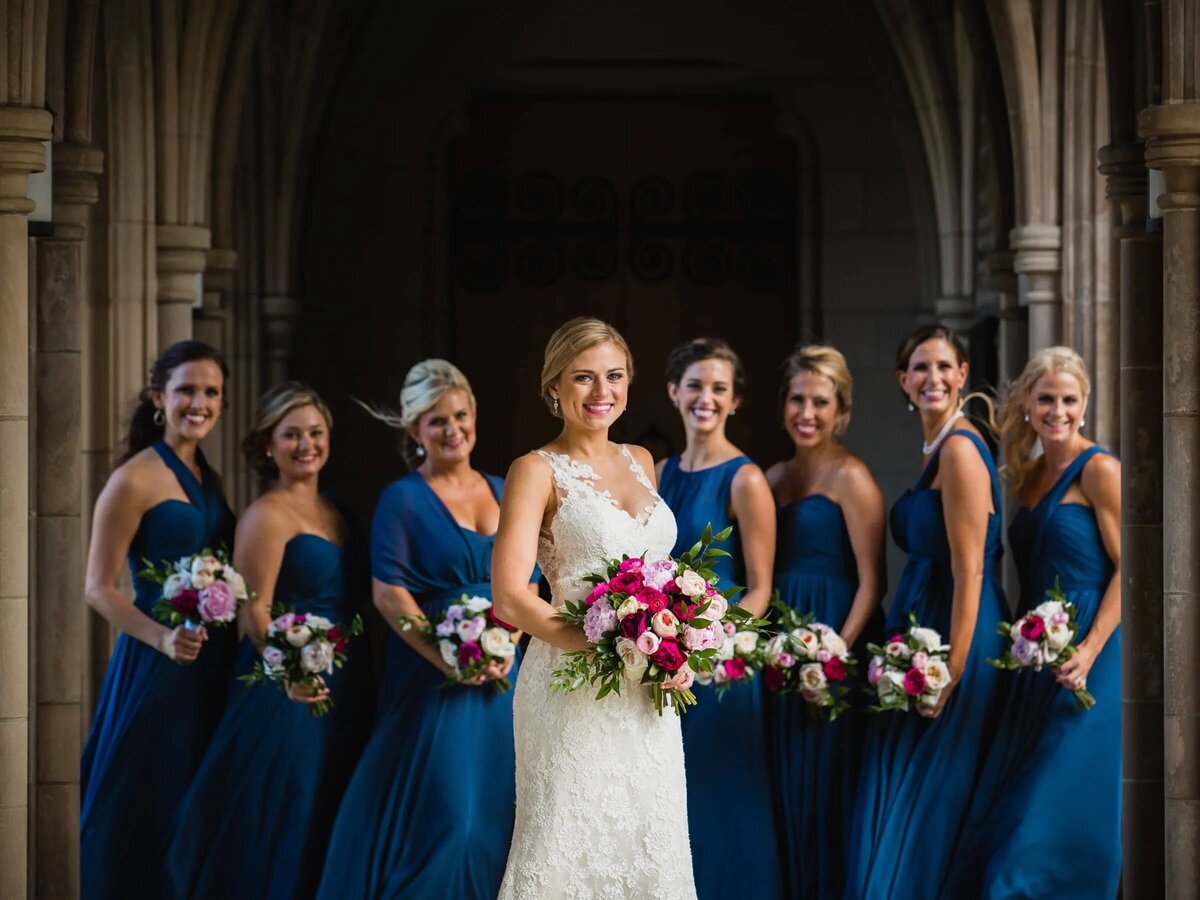 Bride in a lace gown with bridesmaids in royal blue dresses, all holding bouquets, posing in a classic architecture setting