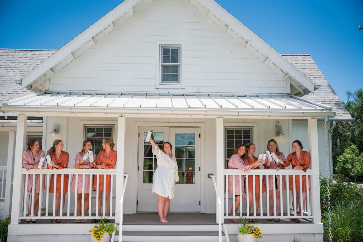 A bride and her bridesmaids stand on the porch of the getting ready cottage at The Barn at Raccoon Creek.
