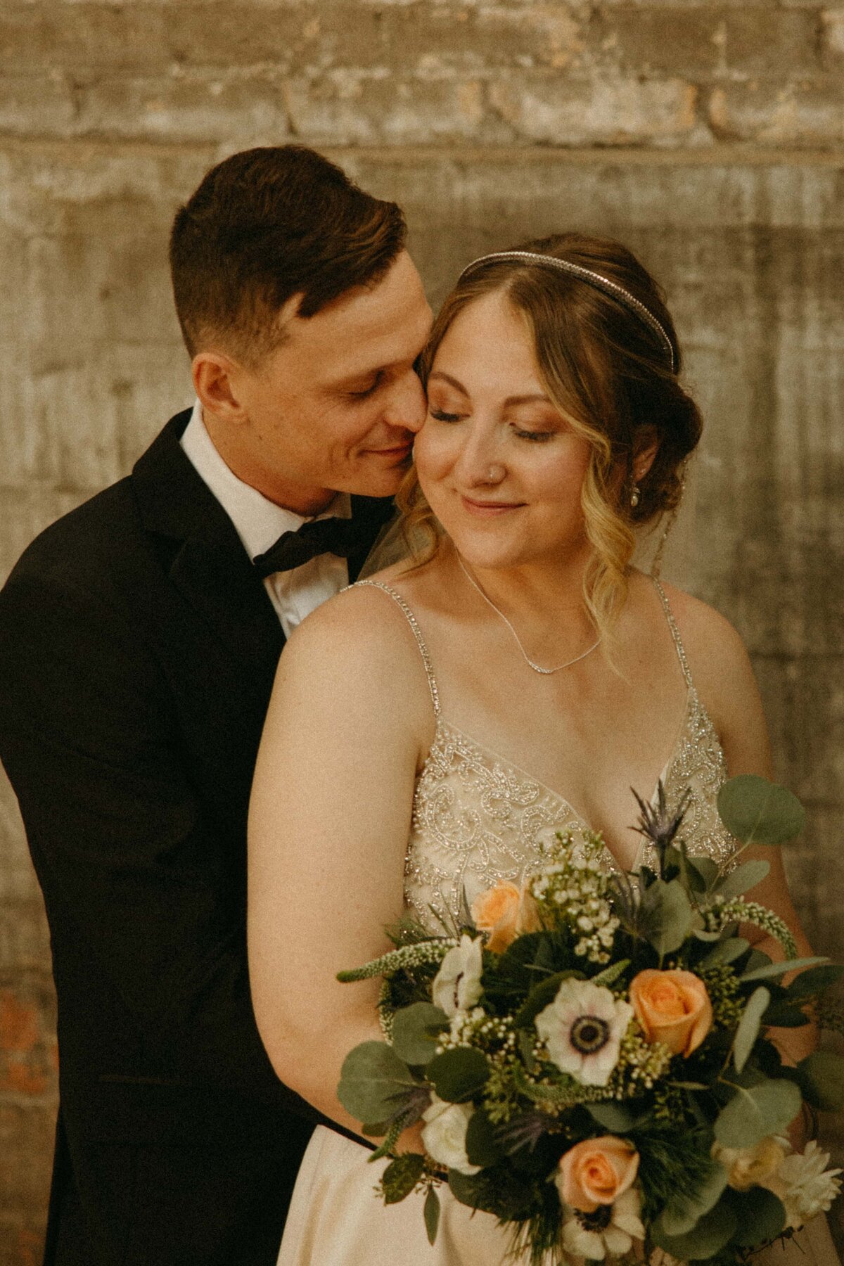 A bride and groom affectionately embracing, with the bride holding a bouquet of flowers, against a rustic brick wall background at Park Farm Winery.