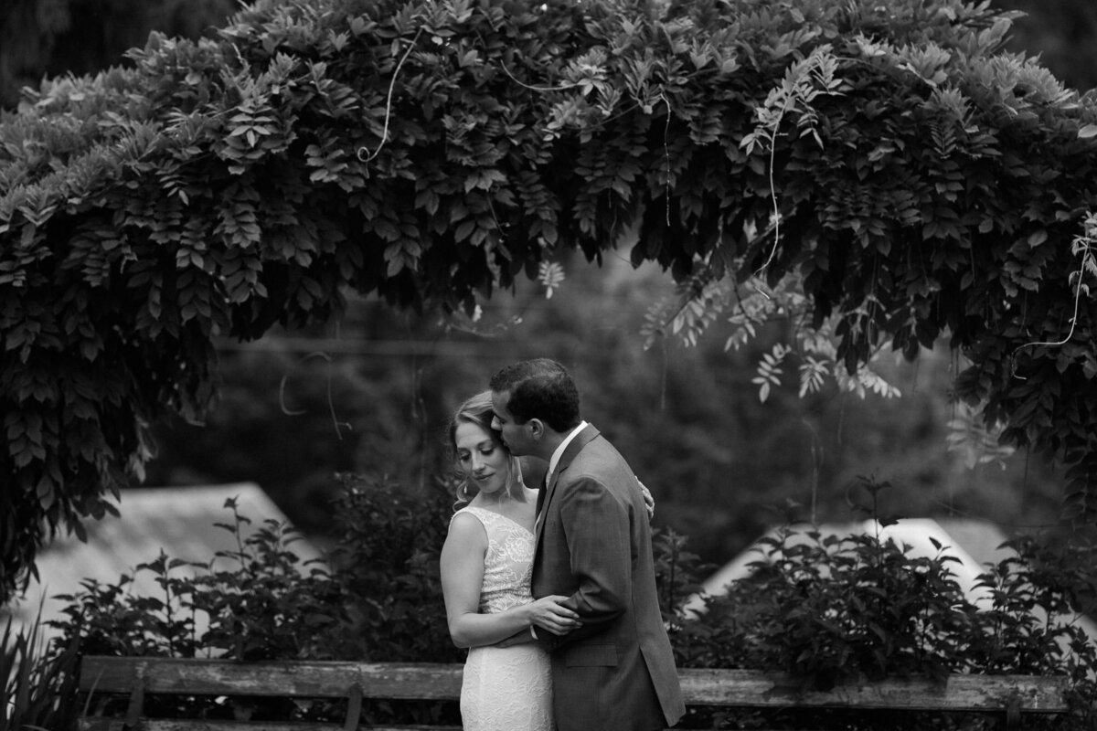 Black-and-white-photo-of-bride-and-groom-kissing-underneath-arbor-at-wedding-venue-Green-gates-at-Flowing-Lake-Snohomish-wedding-venue-photo-by-Joanna-Monger-Photography