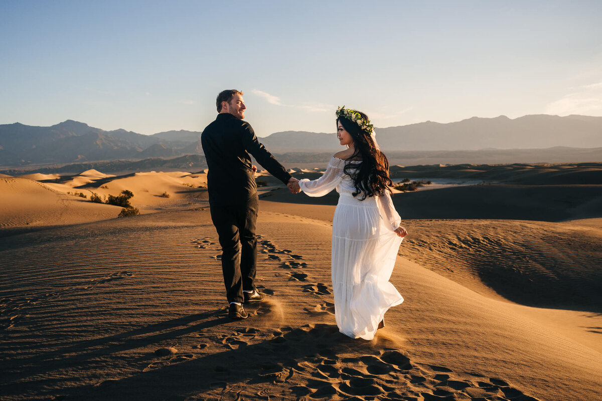 An enchanting elopement photo at Mesquite Sand Dunes in Death Valley National Park, capturing the couple's happiness as they walk hand in hand on the sandy dunes, surrounded by the desert's beauty.
