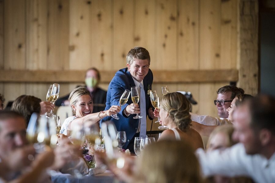 A groom stands and reaches across the table to cheers a champagne glass with his wedding guests.