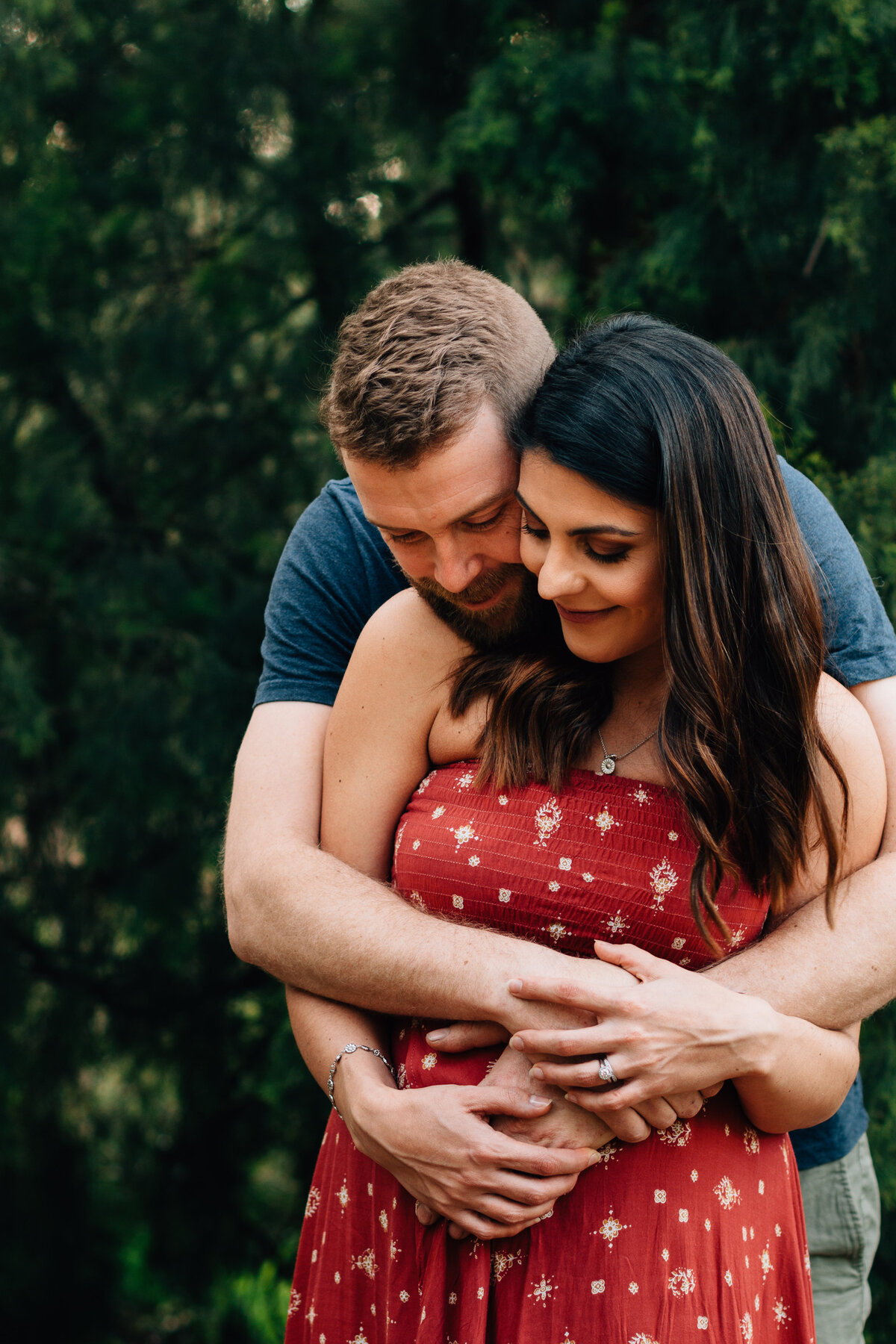 husband hugging wife around pregnant belly outdoors in front of trees
