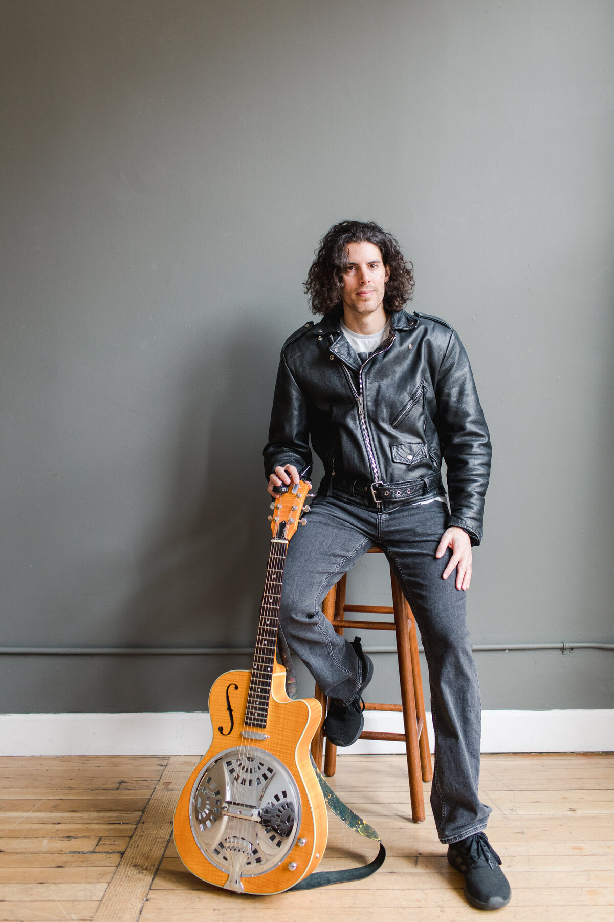 a curly haired man sitting on a stool and a guitar in one hand resting  in front of him