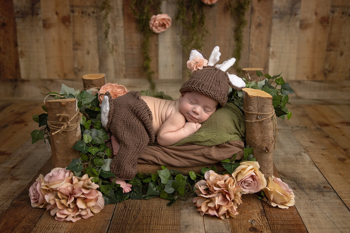 A sleeping baby dressed in a crochet deer outfit rests on a small, rustic wooden bed. The serene scene, crafted by a Jacksonville baby photographer, is adorned with pink roses, greenery, and wooden logs against a backdrop of hanging flowers and lush greenery.