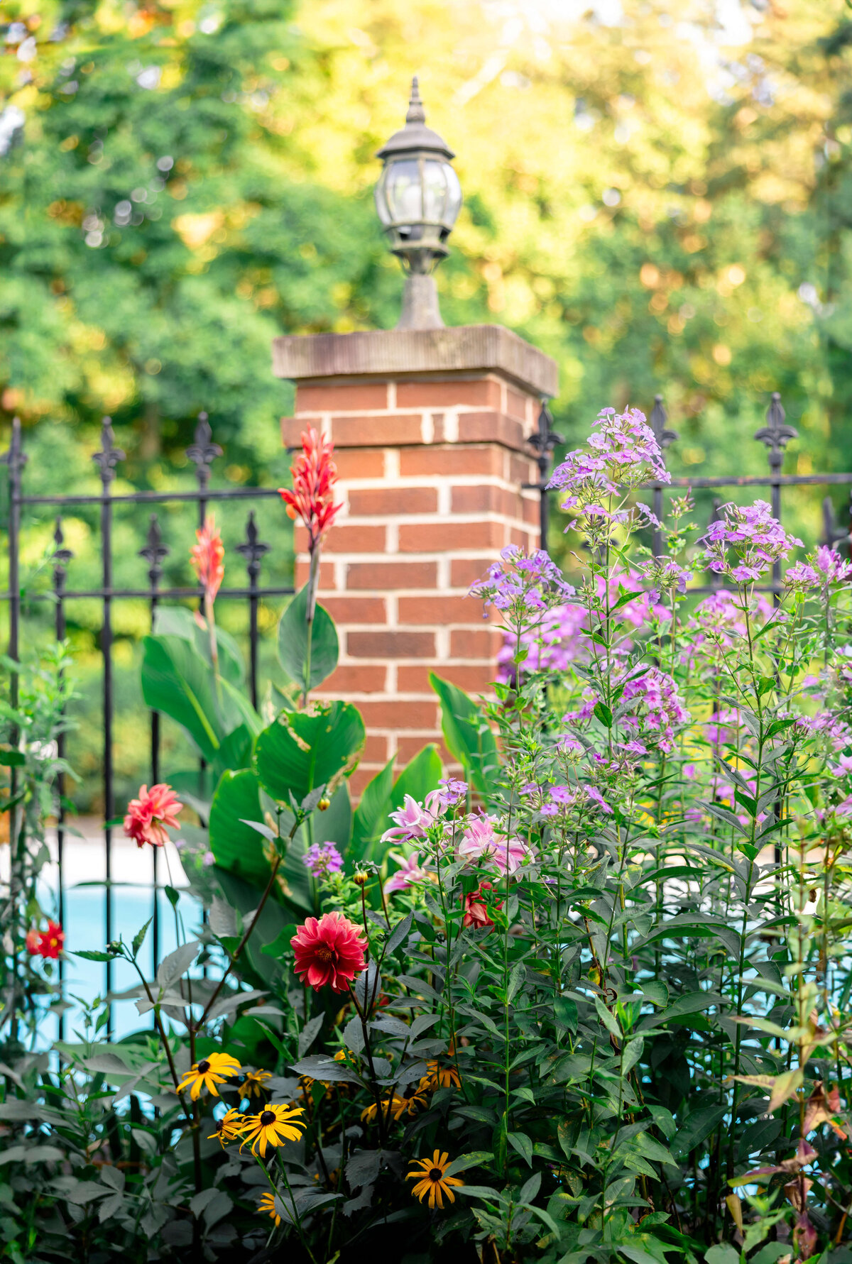 A vibrant garden with various colorful flowers, including pink, red, and yellow blooms, stands in front of a brick column topped with a lantern. A black metal fence borders the garden, and lush greenery is visible in the background.